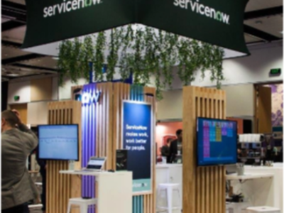 ServiceNow stand with screens on timber-slat pillars under a square hanging banner with greenery