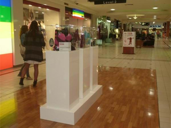 Three tall white plinths on a shared white base in a shopping centre walkway, each topped with a clear showcase holding products