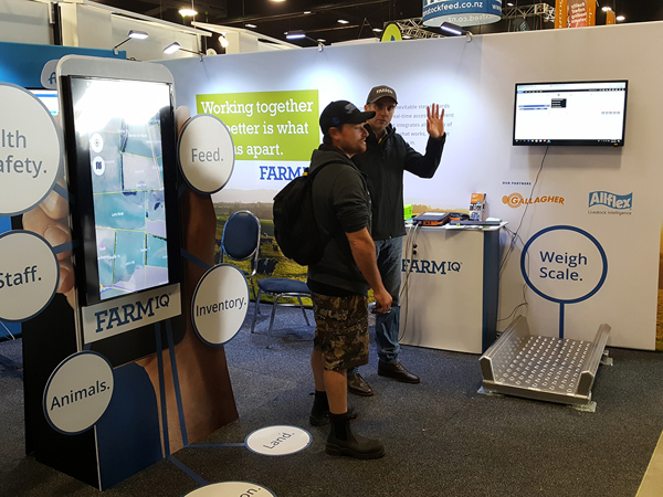 Exhibition stand with a tall phone-shaped touchscreen kiosk showing a farm map, while a staff member talks a visitor through the software beside a wall-mounted screen