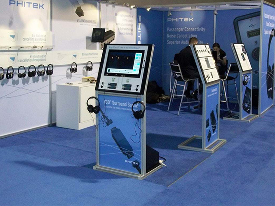 Phitek exhibition stand with three free-standing screen kiosks, headphones for product demonstrations and two people seated on high chairs at the back