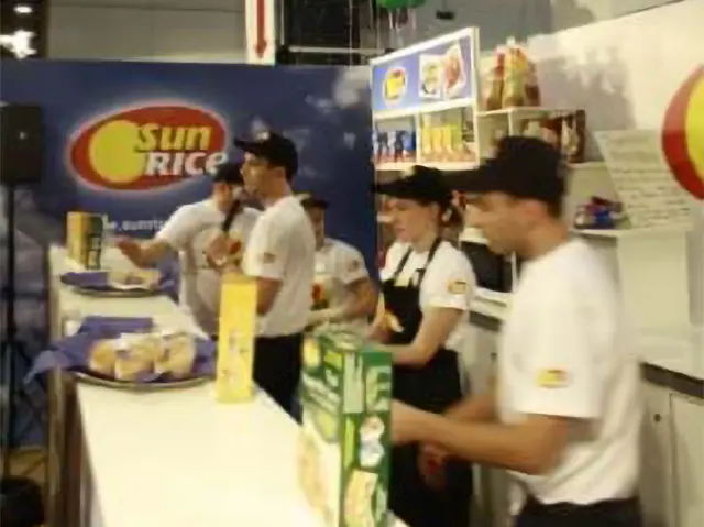 SunRice staff serving food samples from a long white exhibition counter at an expo