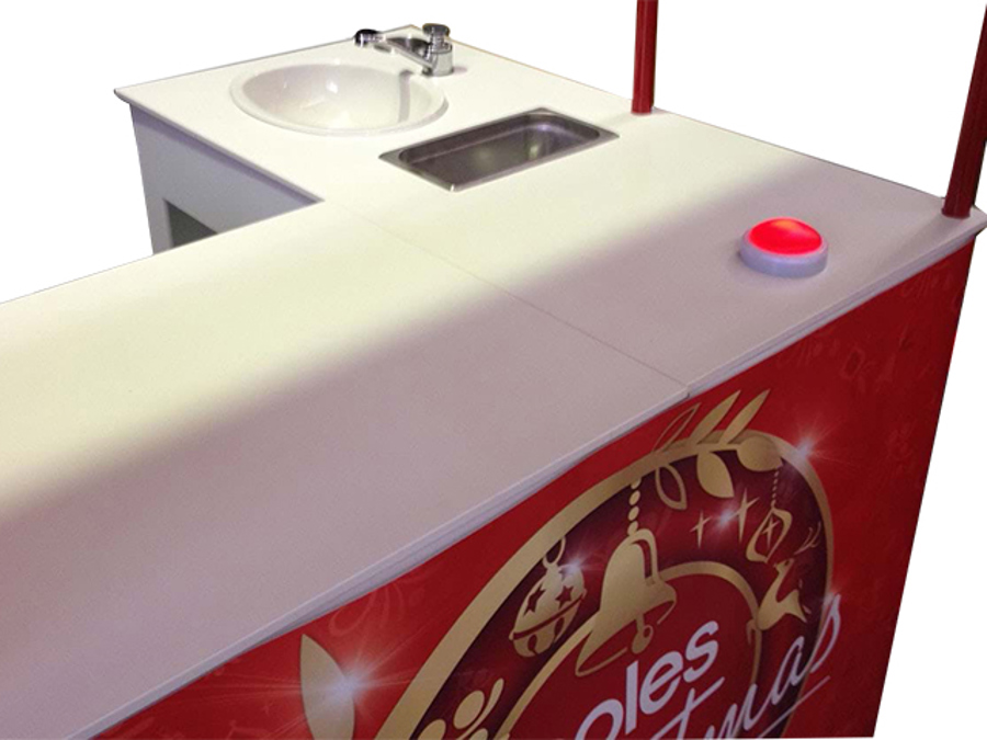 White L-shaped demonstration counter with a small sink and tap, a stainless steel well and red Coles Christmas graphics on the front