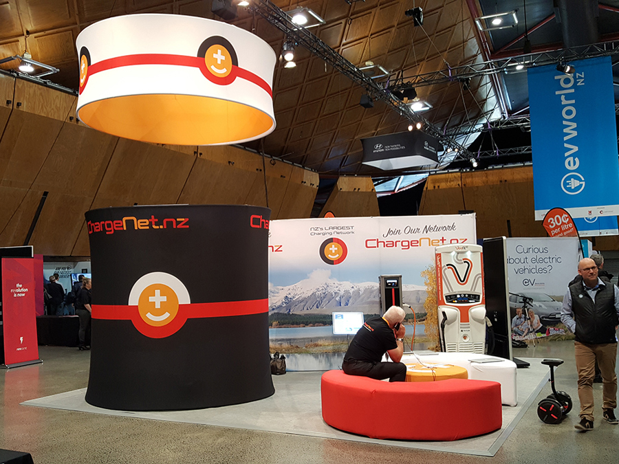 Exhibition stand in a hall with a large round fabric banner hung above it, a tall black fabric cylinder, a printed back wall and red curved seating